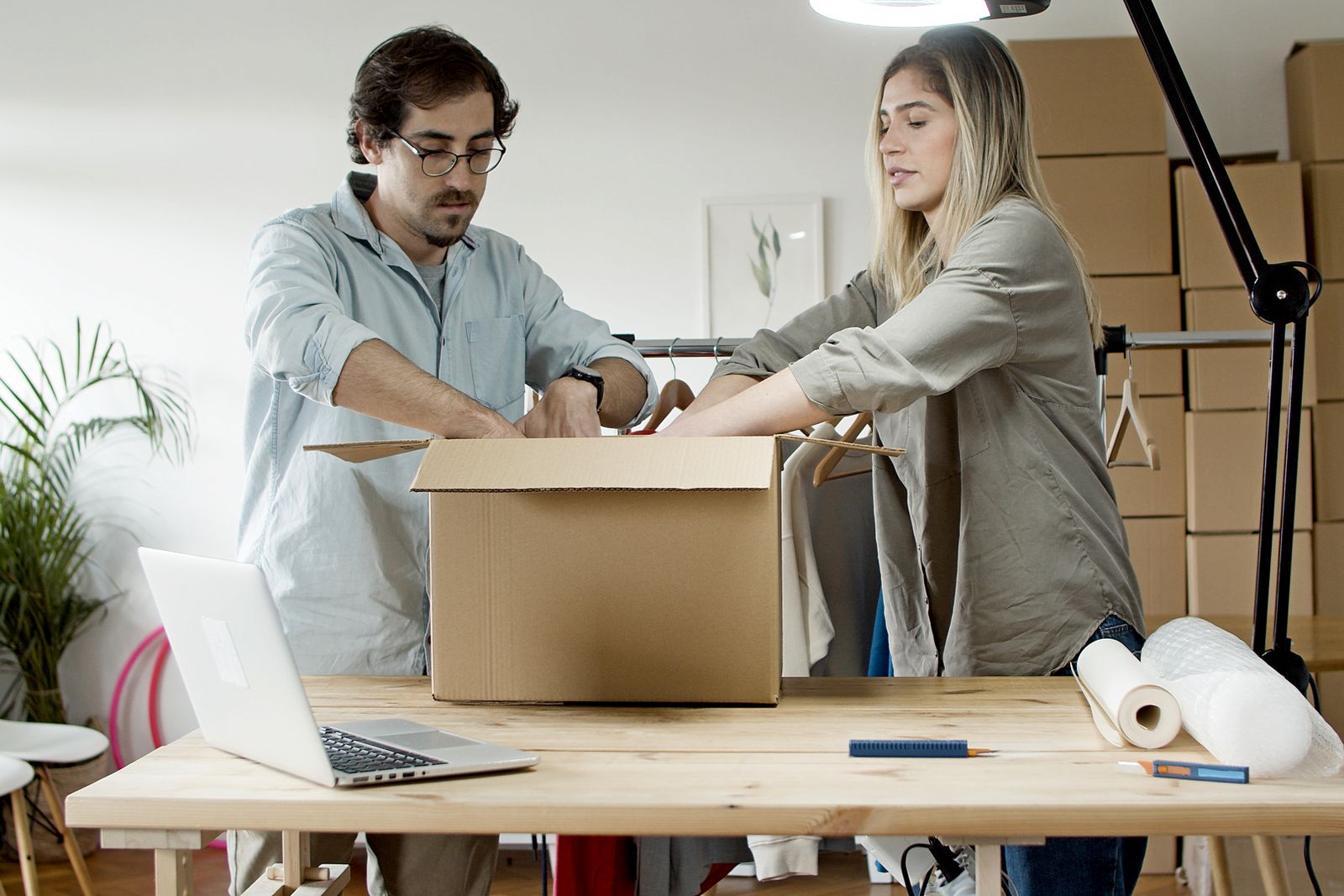 A man and a woman packing ecommerce orders into cardboard boxes at a table with a laptop open, showing the fulfilment and returns details an AI agent checks before recommending a product.