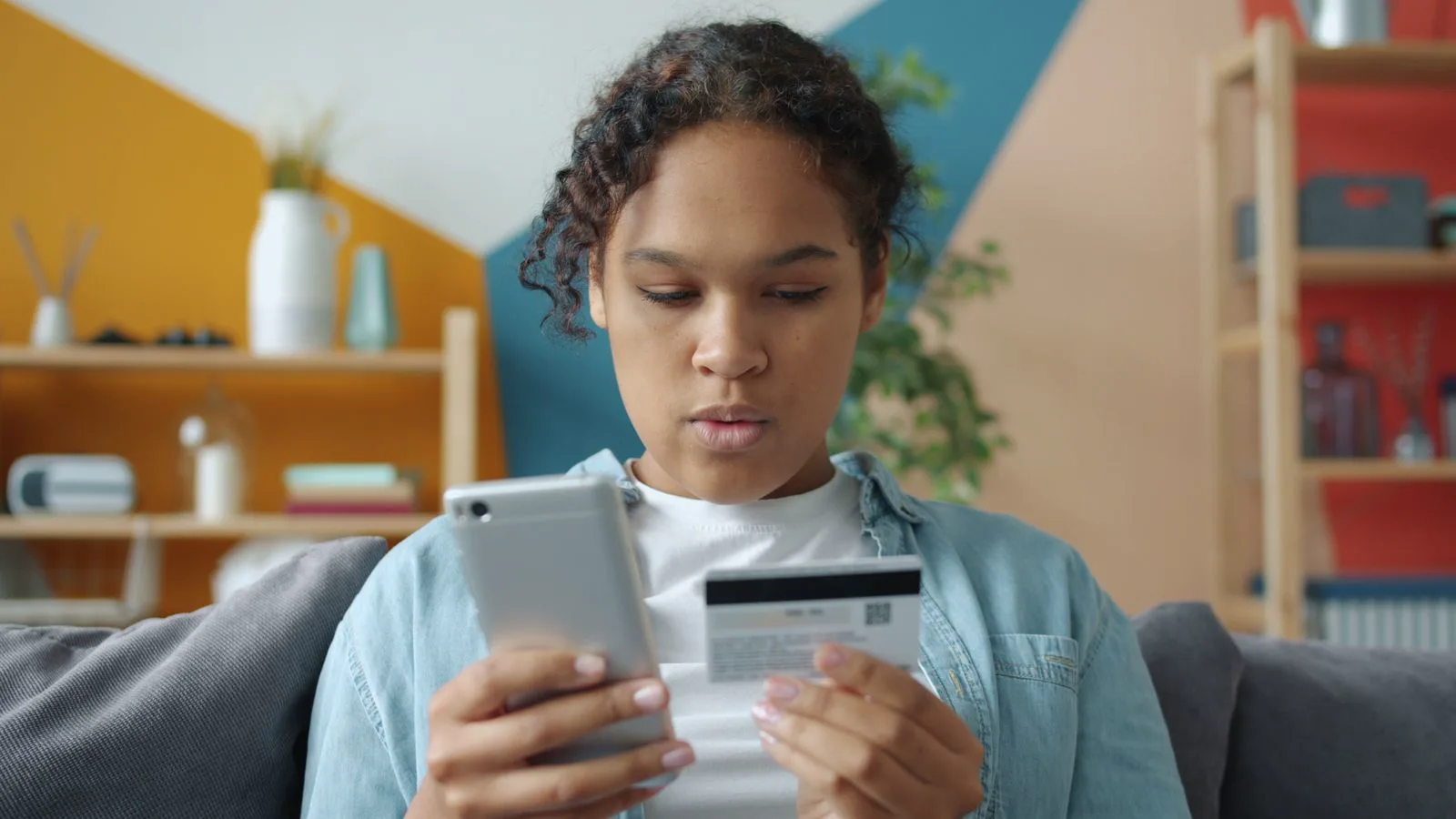 A young woman sitting on a sofa at home, holding a smartphone in one hand and a payment card in the other, representing a shopper buying through an AI shopping assistant.