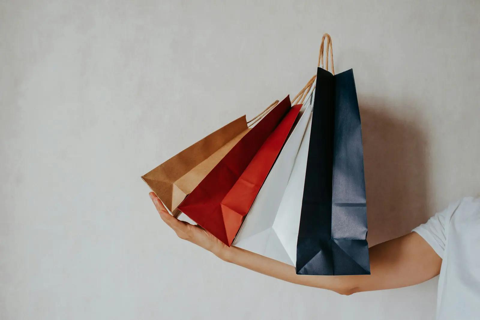 Arm holding out brown, red, white and navy paper shopping bags against a plain wall, representing Black Friday and Q4 retail demand.