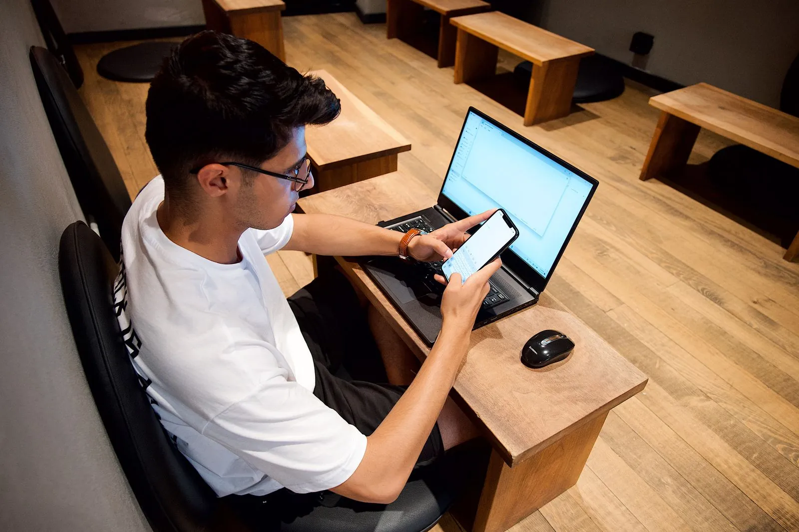 A young man at a wooden desk checking his smartphone with a laptop open in front of him, representing a UK marketer reviewing a brand's Bluesky account.
