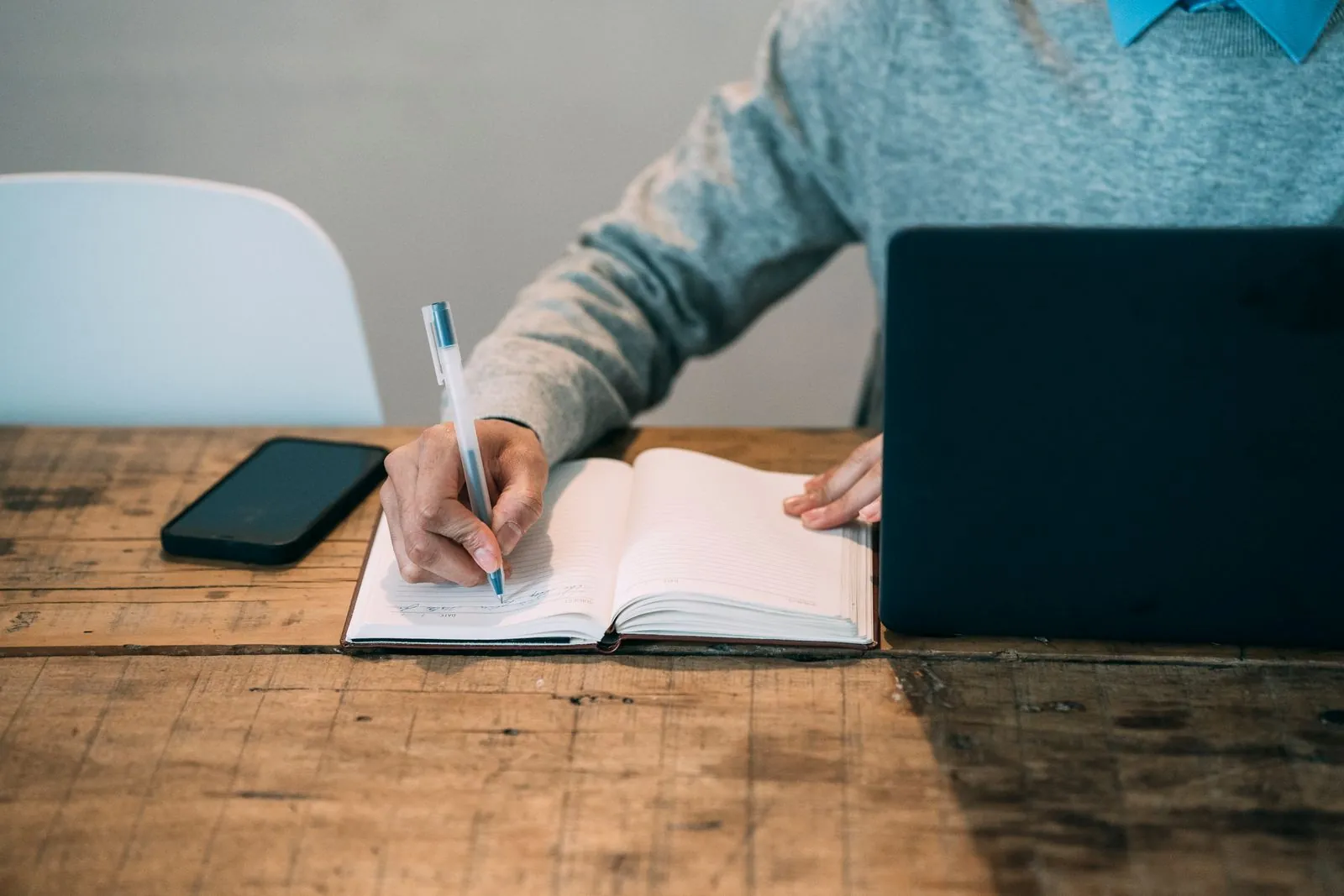 A person writing notes in an open planner beside a laptop and a smartphone on a wooden table, planning a Bluesky content calendar for a UK business.