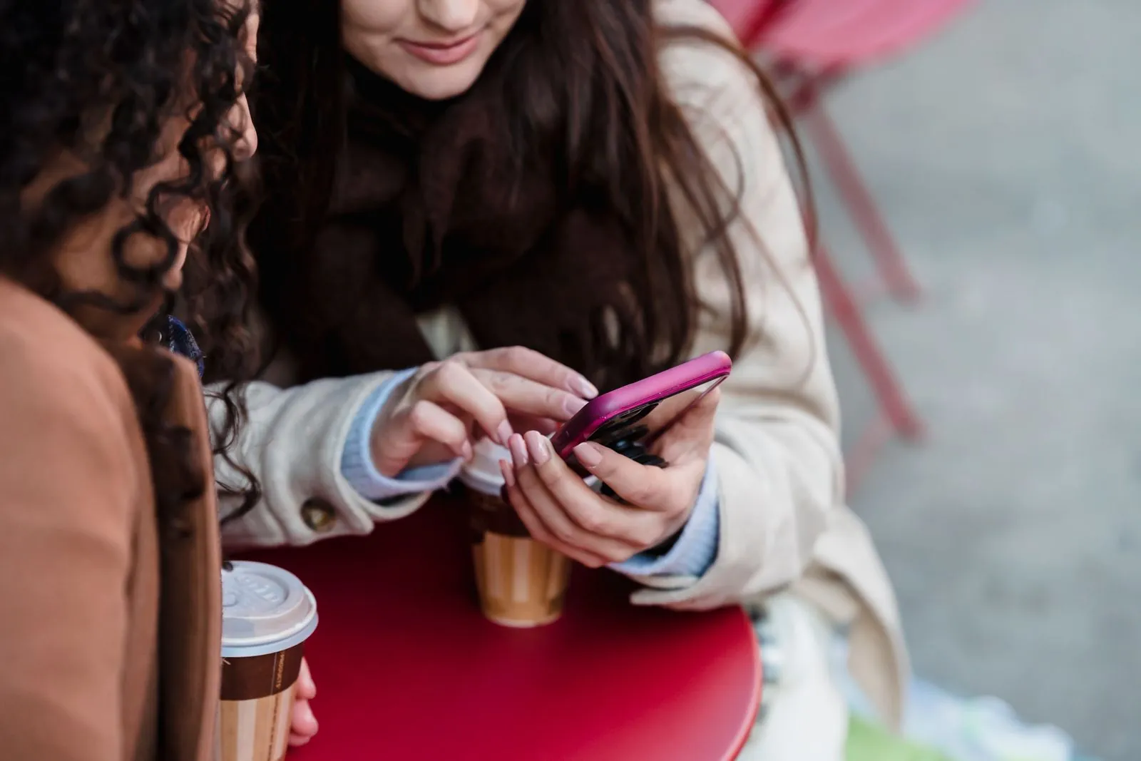 Two friends at a red café table with takeaway coffees, one scrolling a pink smartphone, representing UK consumers browsing a social app like Bluesky.
