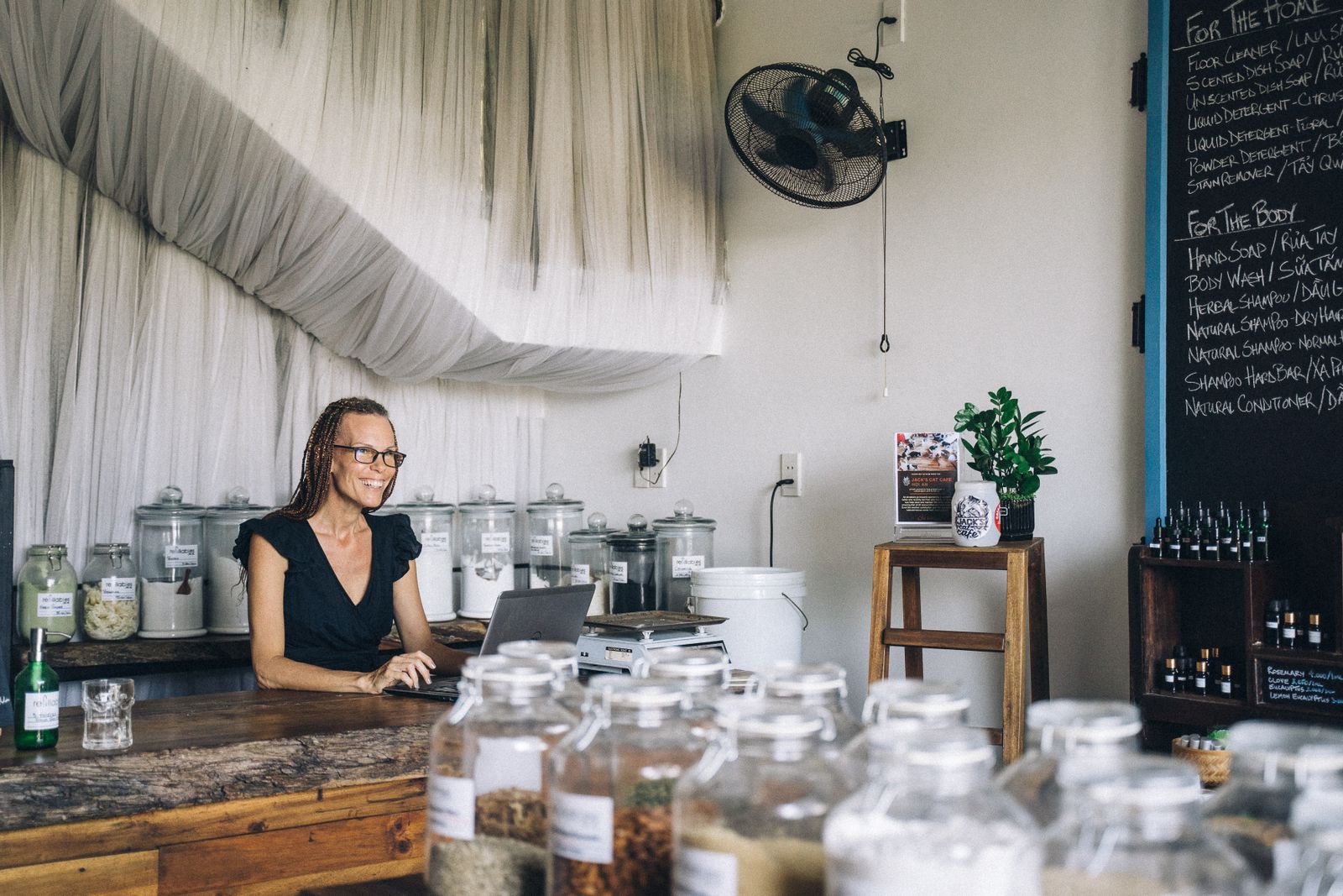 Smiling shop owner working on a laptop at a wooden counter in a small refill shop lined with glass jars, representing a small business checking its online reviews.