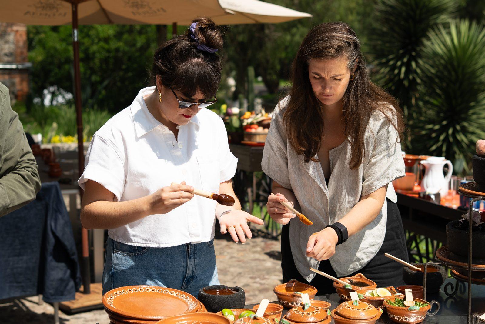 Laura and Andrea from Masafina tasting sauces from small clay pots at an outdoor table.