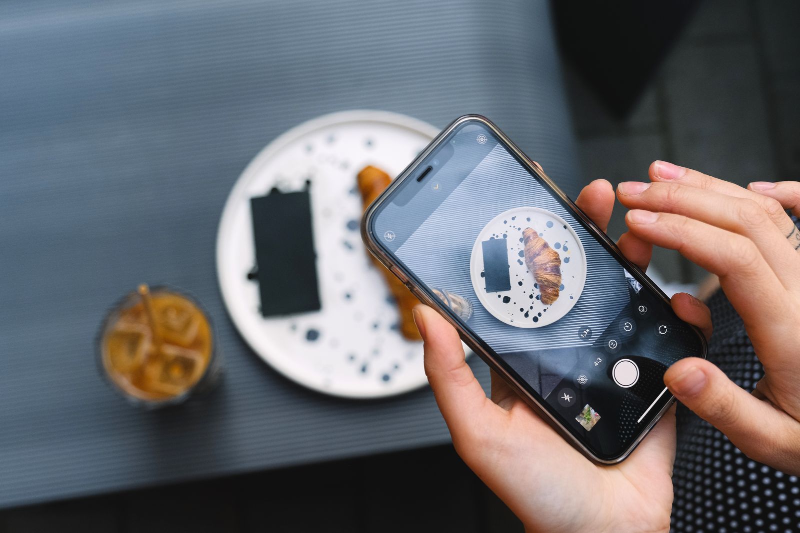 A person holding a smartphone over a café table, photographing a croissant on a speckled plate next to an iced coffee, the kind of hosted food content now covered by UK rules on gifted collaborations.