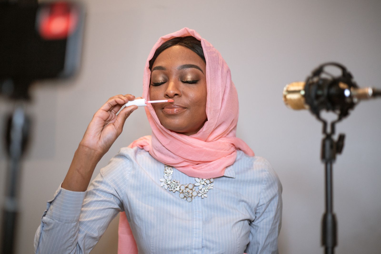 A creator in a pink headscarf testing a lip product in front of a camera and microphone in a home studio, deciding whether a gifted collaboration is worth the time it takes to film.