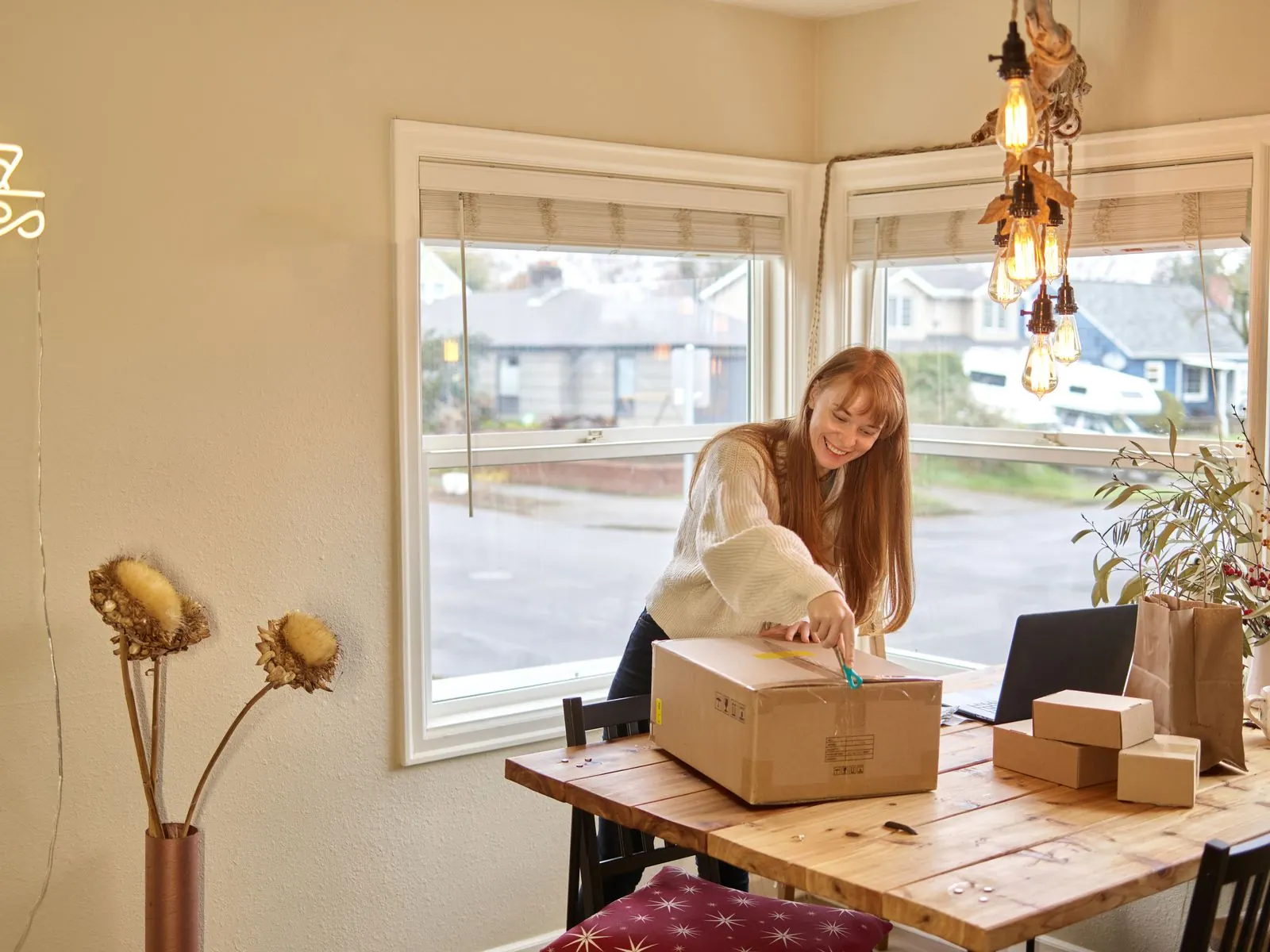 A smiling creator opening a cardboard parcel on a wooden table beside a laptop and smaller boxes, representing a UK creator unpacking a PR package sent as a gifted collaboration.