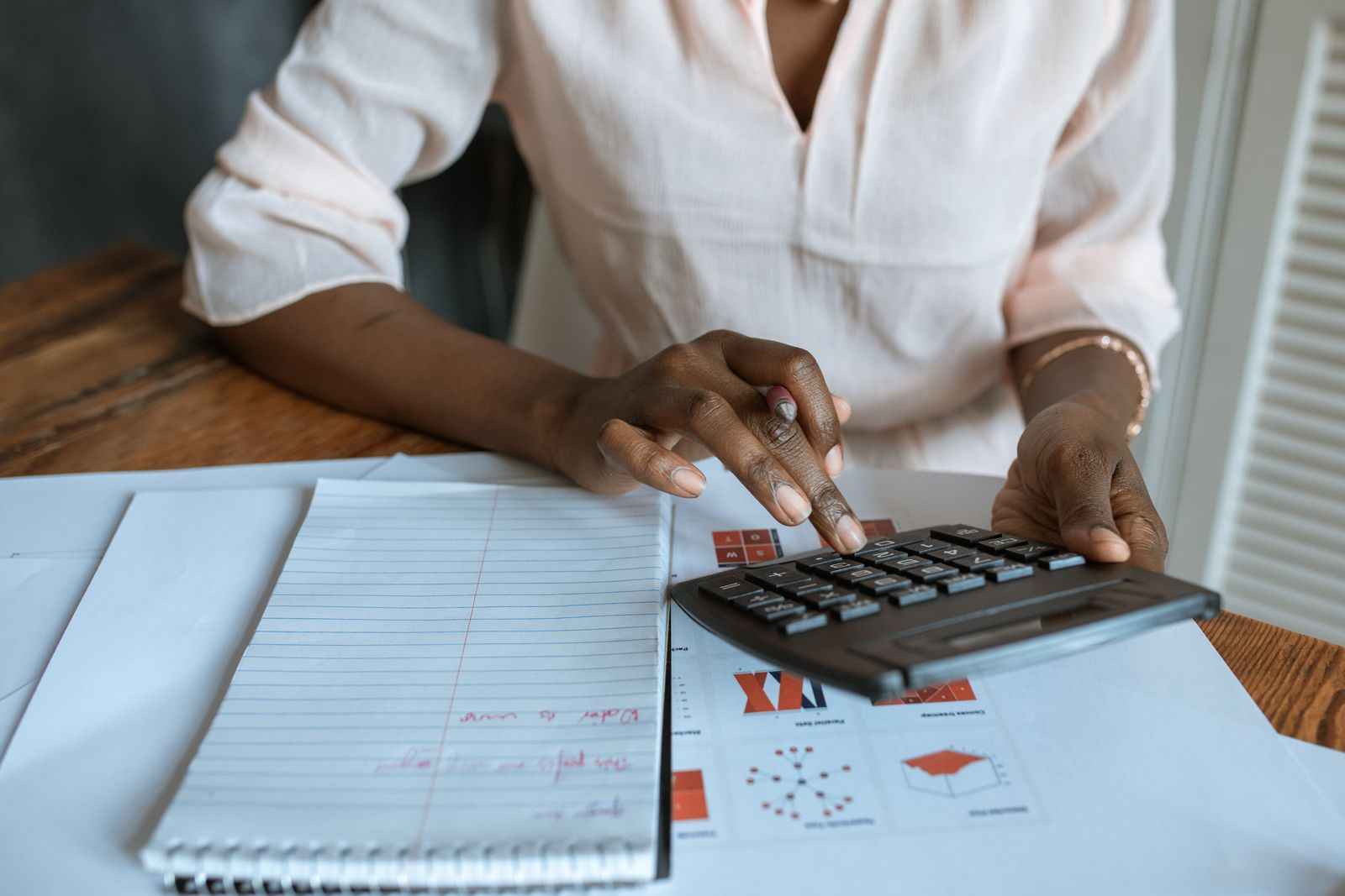 A woman using a calculator at a desk covered with notebooks and printed charts, representing an advertiser working out how much daily budget to allocate to low priority Google Ads channels.
