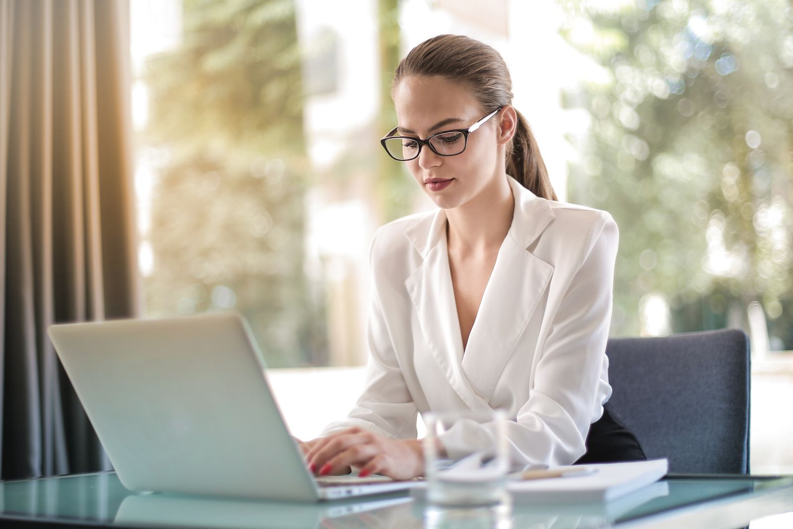 A businesswoman in glasses and a white blouse typing on a laptop at a glass desk in a bright office, representing an advertiser reviewing Google Ads channel settings.