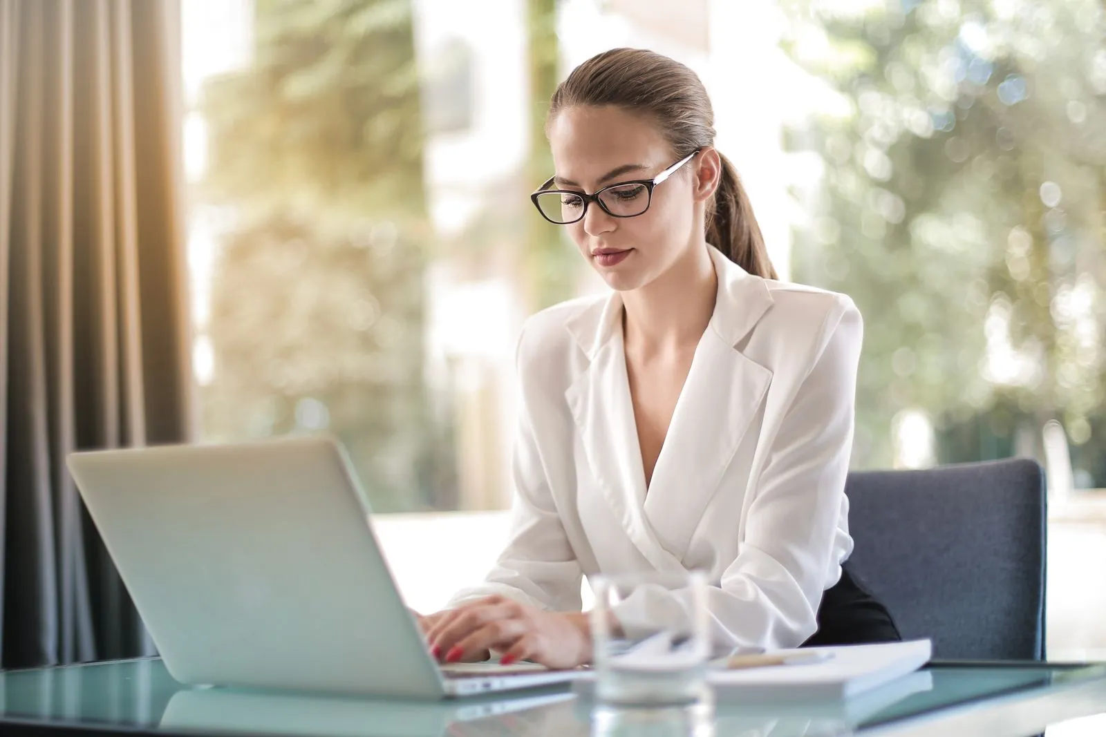 A businesswoman in glasses and a white blouse typing on a laptop at a glass desk in a bright office, representing an advertiser reviewing Google Ads channel settings.