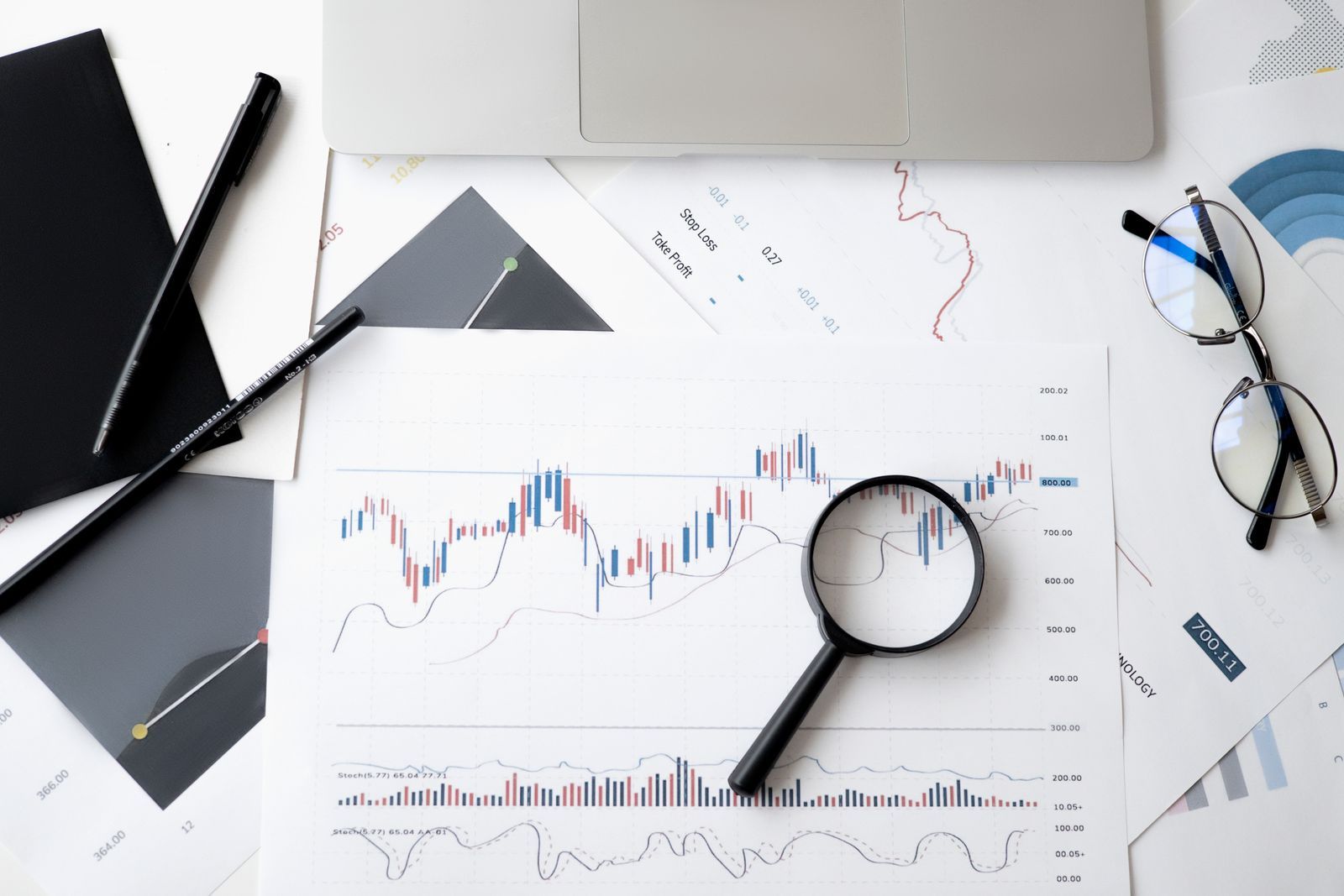 A magnifying glass lying on printed line and candlestick charts next to pens, glasses and a laptop, a reminder to compare rank tracker data with Search Console and real sales before deciding anything.