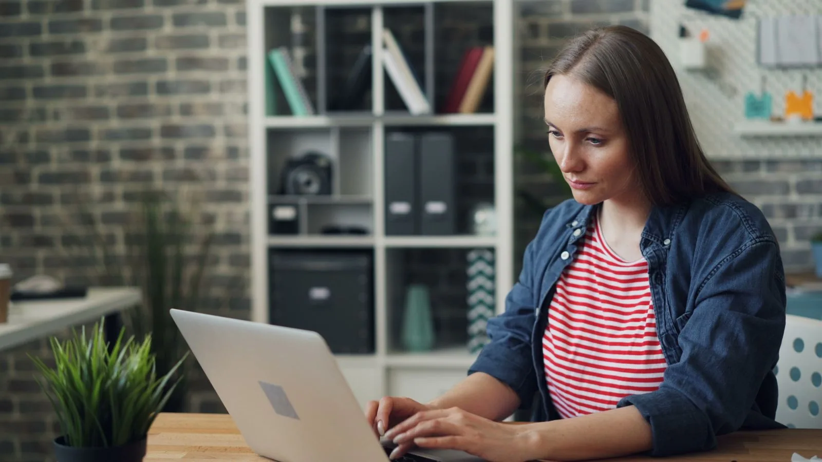 A woman in a striped top and denim shirt working on a laptop at a desk in an office with exposed brick walls, checking Search Console to see whether the Google spam update caused a drop in rankings.