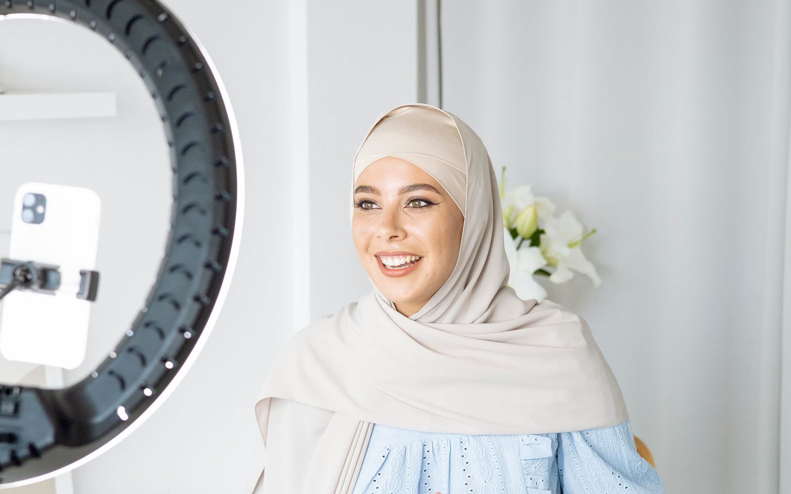 A creator sitting beside a ring light with a smartphone mounted in it, smiling as she talks to camera in a bright home studio, the kind of setup brands look for in a creator media kit.