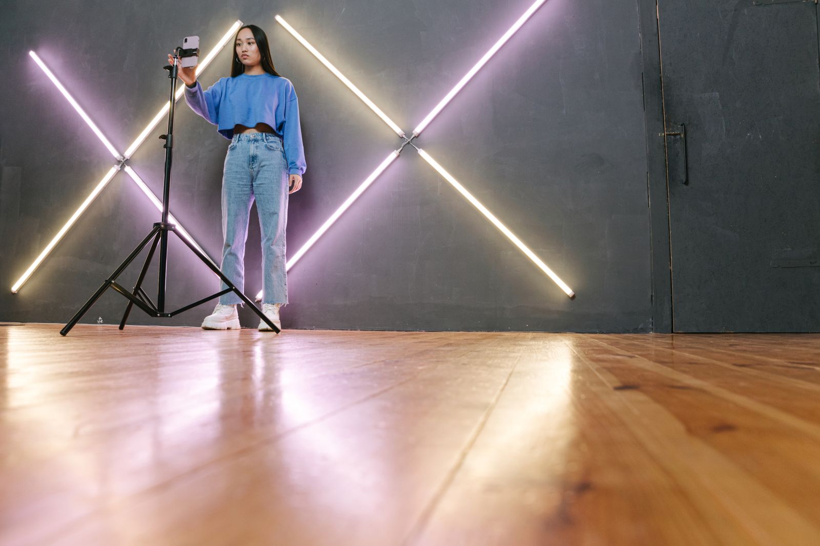 A young creator in a studio lined with neon light bars, framing a shot on a smartphone mounted on a tripod, representing a UK creator producing content for brand deals.
