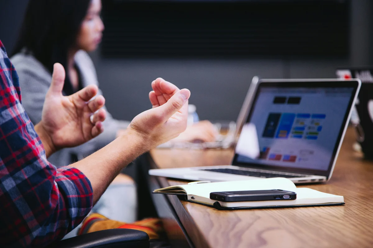 Two colleagues in a meeting, one gesturing while discussing data shown on a laptop screen