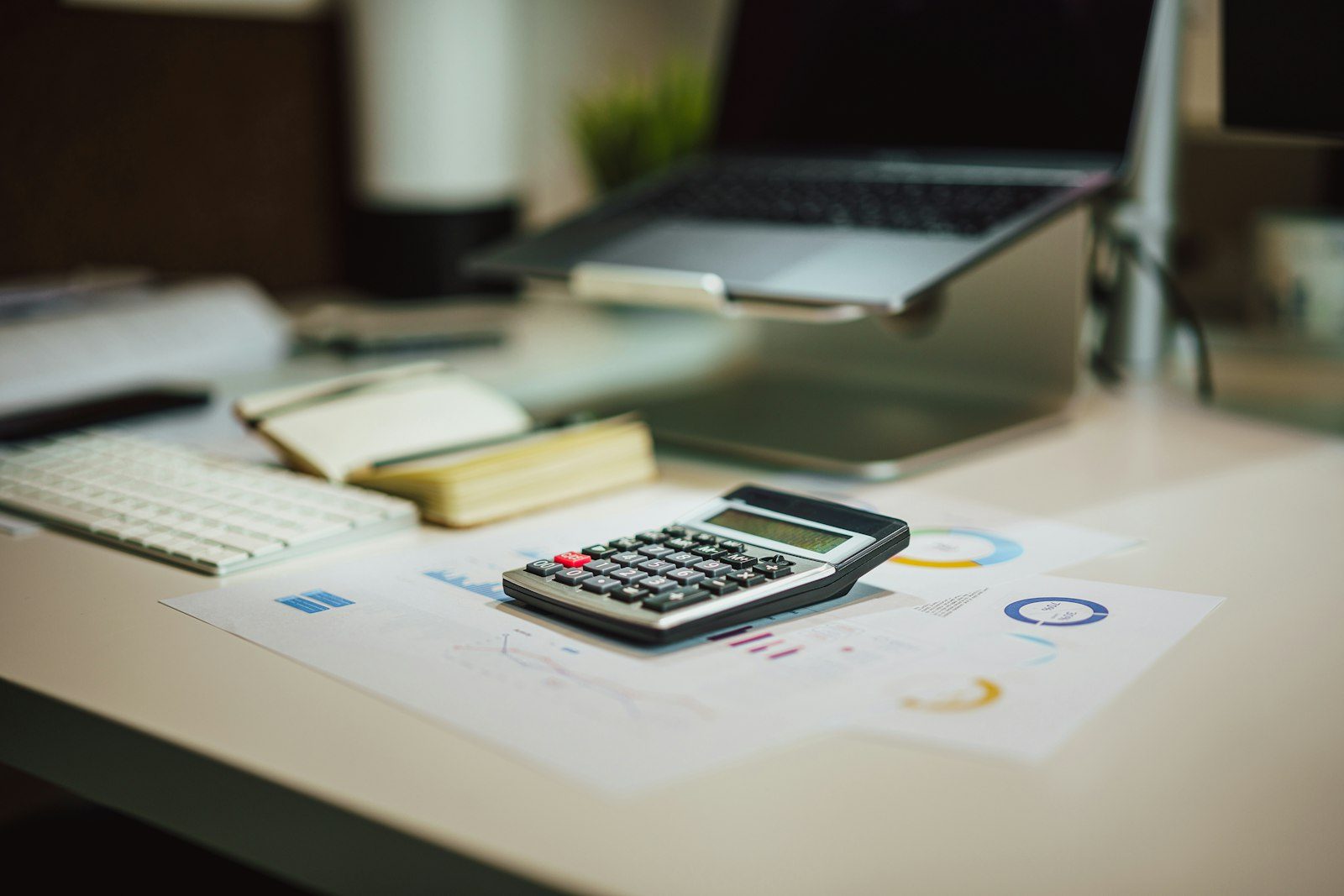 Calculator on printed charts beside a laptop, notebook and keyboard on a desk, representing a creator checking invoices and payment terms.