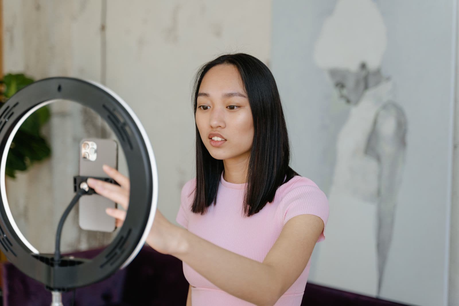 A young woman adjusting a smartphone mounted in a ring light while filming social media content at home.