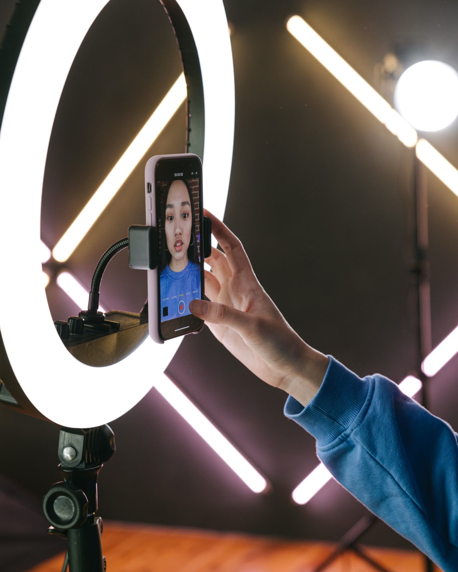 A woman filming herself on a smartphone held in a ring light in a dark studio with neon light tubes behind her