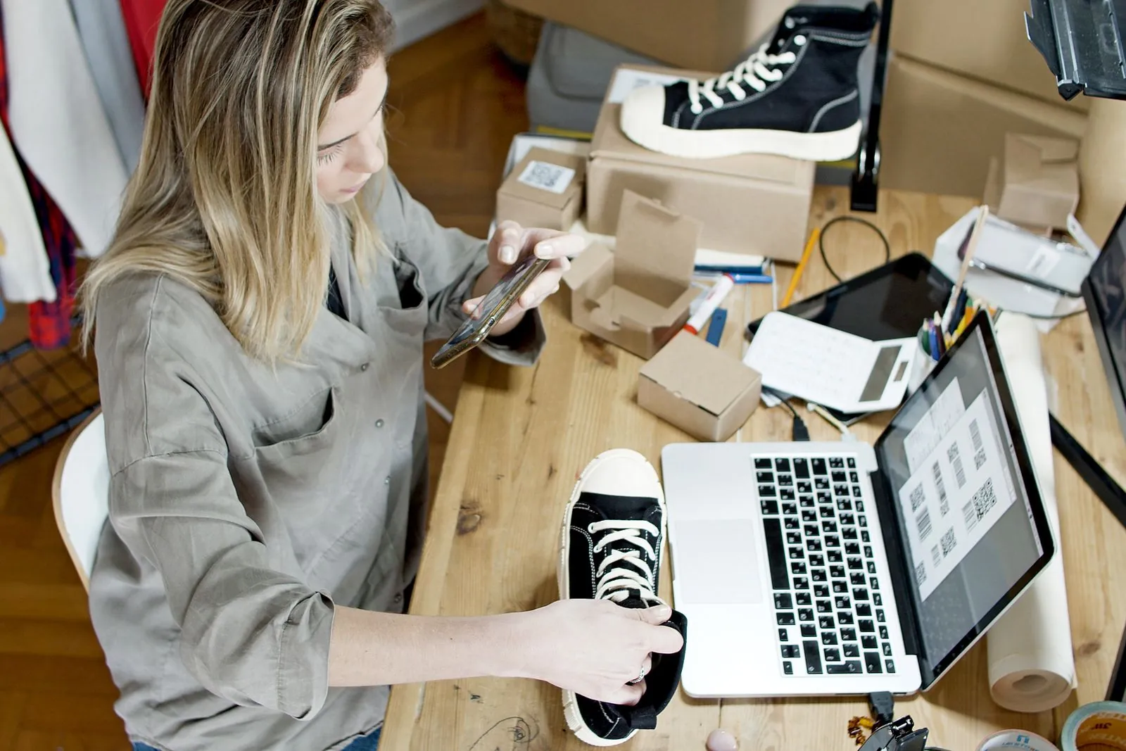 A small business owner photographing a trainer with her smartphone at a desk covered in parcels and a laptop, preparing a social post for Facebook or Instagram.