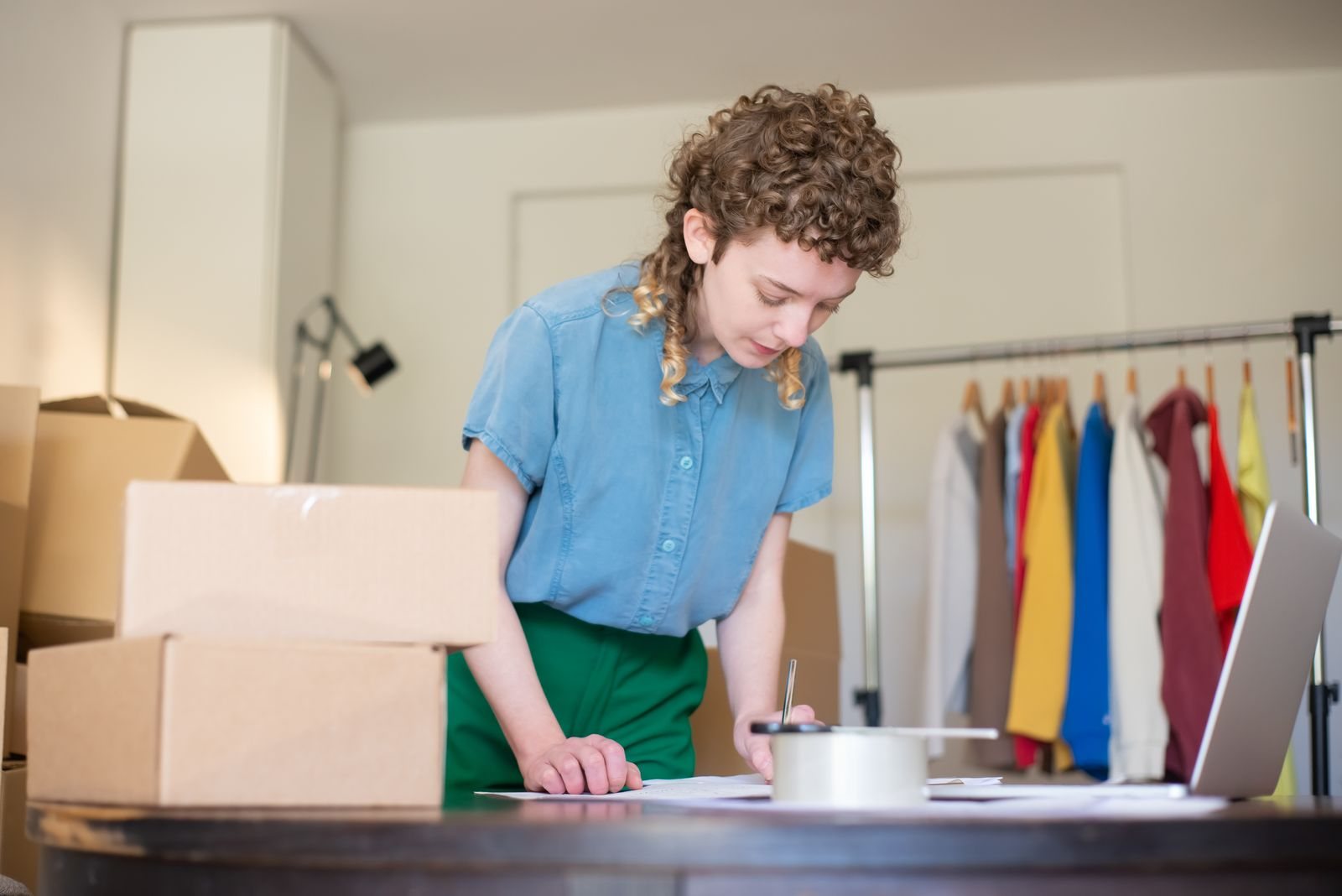 A woman packing online orders beside cardboard boxes, a laptop and a clothing rail, representing a UK ecommerce business deciding whether Meta product browsing suits its catalogue.