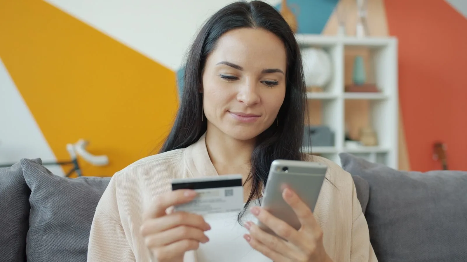 A woman sitting on a sofa holding a smartphone and a bank card, representing a shopper tapping a Facebook or Instagram ad with Meta product browsing switched on.