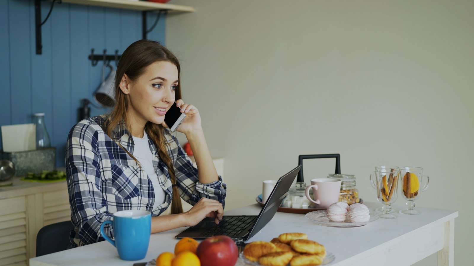 Woman working on a laptop while taking a phone call at a bright kitchen table, standing in for UK SME owners researching and responding across devices.