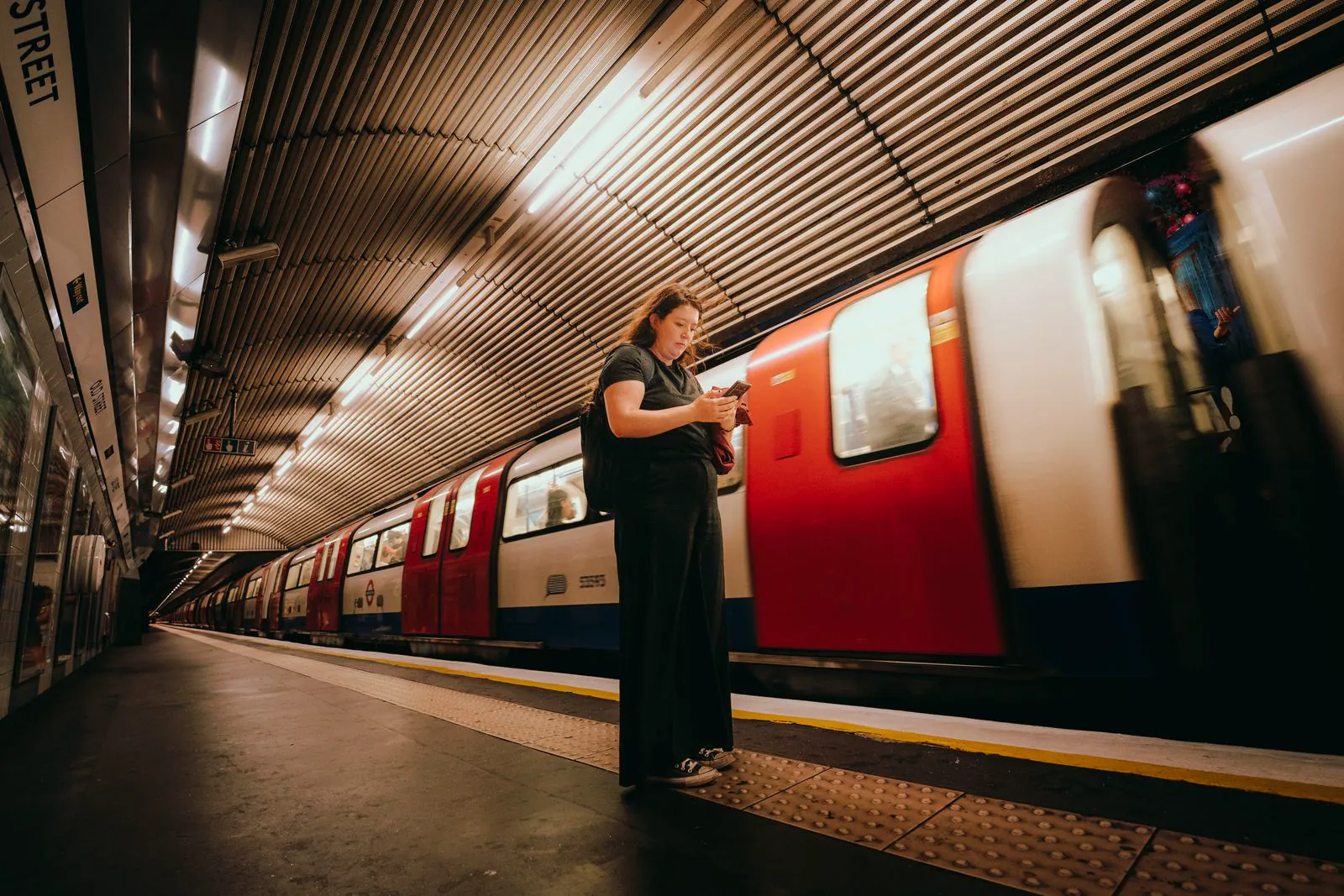 A woman checking her phone on a London Underground platform as a red and white Tube train pulls in, representing UK audiences scrolling Threads on the move.