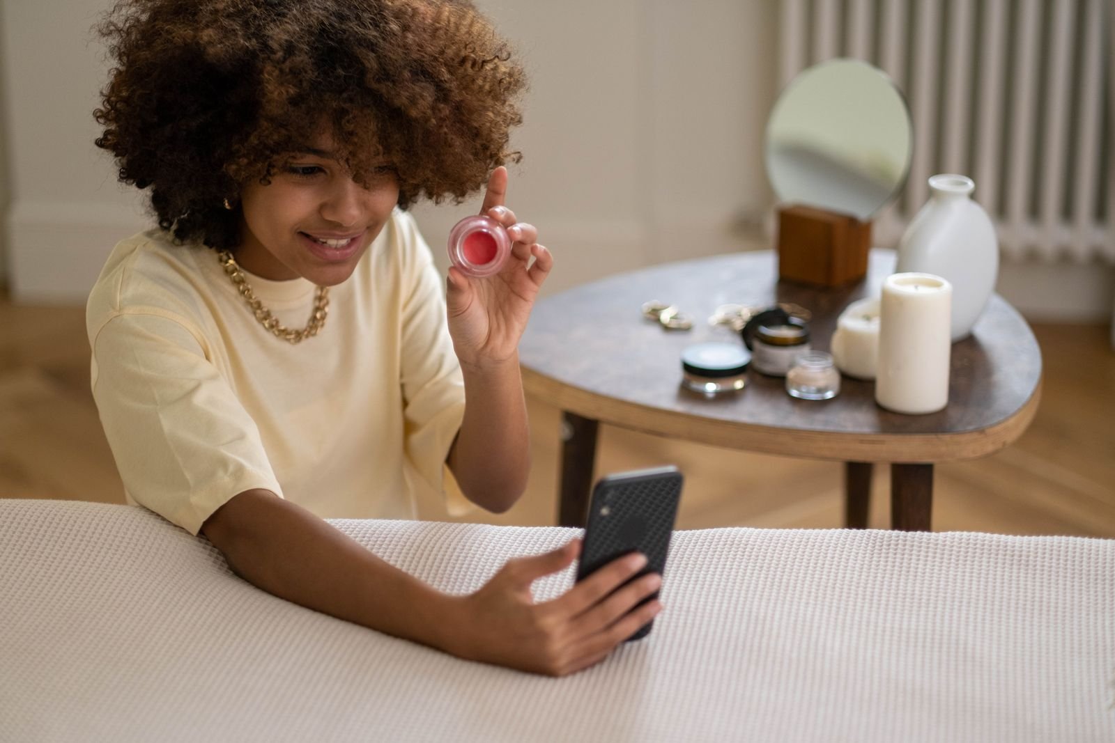 A smiling creator in a pale yellow T-shirt holding up a small pot of lip and cheek tint while filming herself on a smartphone, the kind of product video UK brands commission from UGC creators.
