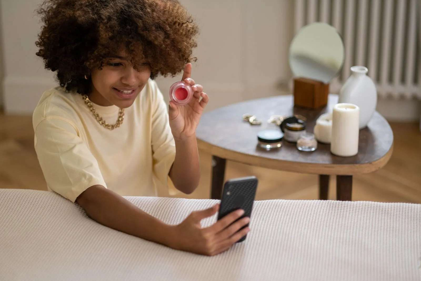 A smiling creator in a pale yellow T-shirt holding up a small pot of lip and cheek tint while filming herself on a smartphone, the kind of product video UK brands commission from UGC creators.