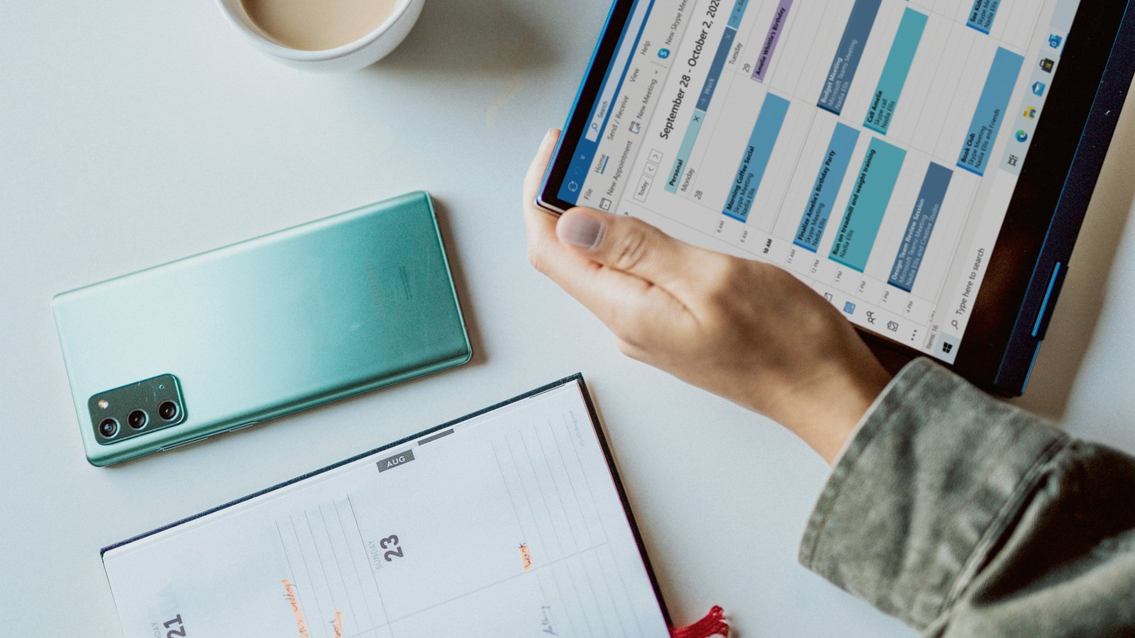 A person holding a tablet showing a calendar app, next to a coffee mug and a paper planner
